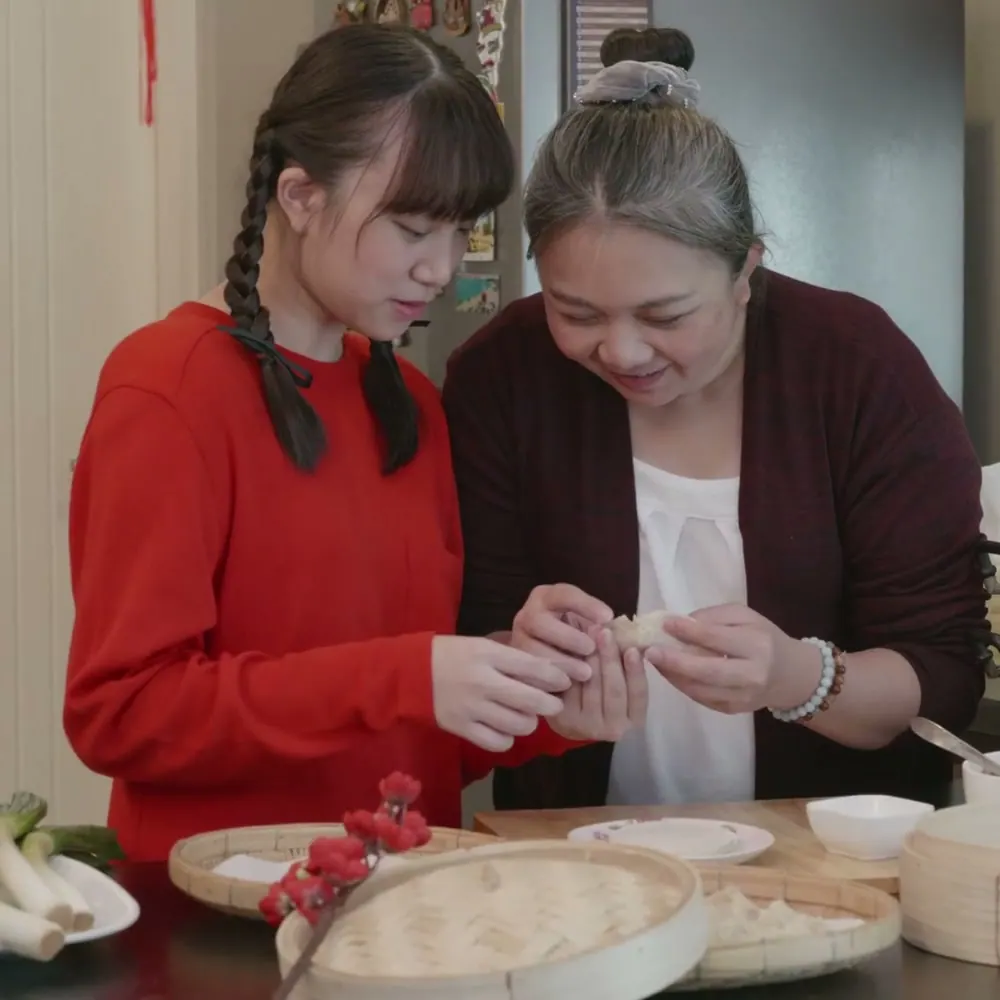 A grandmother and her granddaughter smile as they fold dumplings together over a steamer basket.